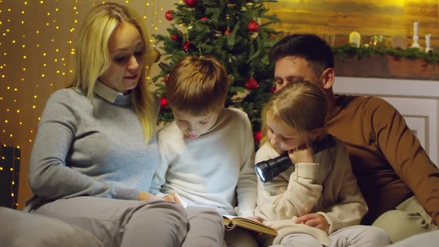 Little boy and girl holding flashlight and reading a book with loving mother and dad while sitting together in cozy living room decorated with fairy lights and Christmas tree