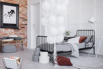 Stylish bedroom interior with grey bedding, bunch of white balloons and map in black frame on the brick wall, real photo