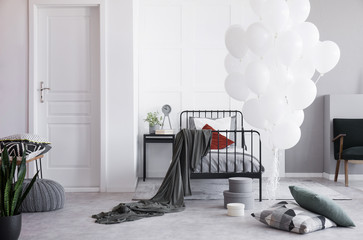 Bunch of white balloons next to single metal bed with grey bedding and white and dark red pillows in bright scandinavian bedroom interior, real photo