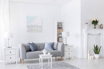 Table on carpet in front of grey settee in white living room interior with lamps and poster. Real photo