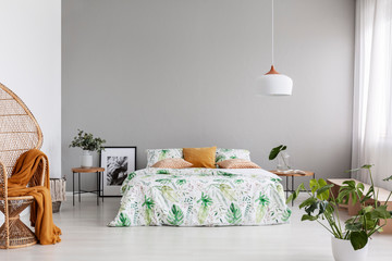 Copy space on empty grey wall of stylish bedroom with leaf patterned bedding on bed and peacock chair in the corner, real photo - obrazy, fototapety, plakaty