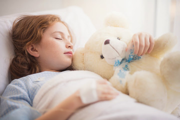 Adorable little girl resting in a hospital bed with her teddy bear. Selective focus at her left eye.