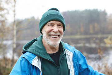 Happy retirement concept. Picture of joyful Caucasian unshaven senior male wearing green hat, hoodie and jacket looking at camera with cheerful broad smile, enjoying warm autumn morning outdoors