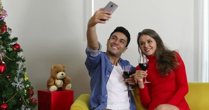 Young good looking Caucasian Couple sitting on sofa and drinking wine together, they selfie a photo by man cellphone, celebrated their special time, concept for Christmas festival.