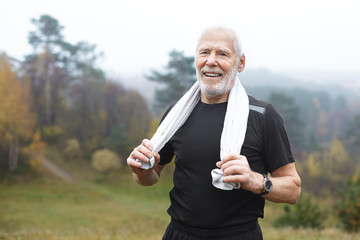 Happy elderly Caucasian male with stubble choosing active lifstyle, smiling broadly after intensive morning cardio workout in park, wiping sweat using white towel. Health, well being and energy - Powered by Adobe
