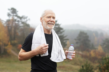Exhausted elderly male with gray hair and beard drinking water after outdoor exercise and wiping...