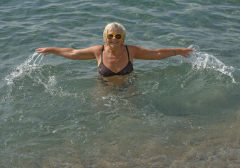 Aged woman is doing splashing motions in clear sea water.