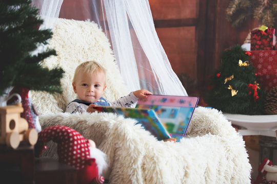 Cute Boy Reading Book Sitting On The Chair Under The Christmas Tree At Home