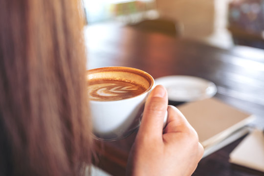 Closeup image of a woman holding and drinking hot coffee with notebooks on table in cafe