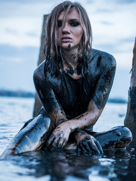 Portrait of Blonde Caucasian Girl Sitting Smeared in a Healthy Black Mud in old Firth with Wooden Posts for Salt Production