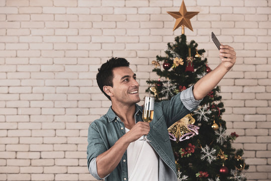 The satisfied caucasian man holding a glass of dazzling white wine is taking his selfie in his special time on christmas. The background of decorated christmas tree with a copyspace. christmas theme