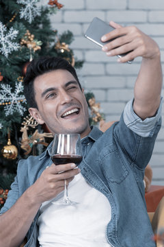 The caucasian man with glass of red wine is taking a selfie with a gorgeous smile on his face in the amazing christmas party inside a room full of christmas trees and decorations. christmas theme.