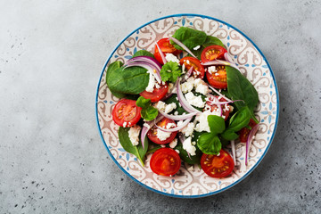 Tomatoes, spinach leaves, red onions and feta cheese salad on light ceramic plate on gray backround. Selective focus. Top view.