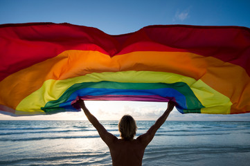 Silhouette of man holding gay pride rainbow flag in front of tropical beach sunrise scene