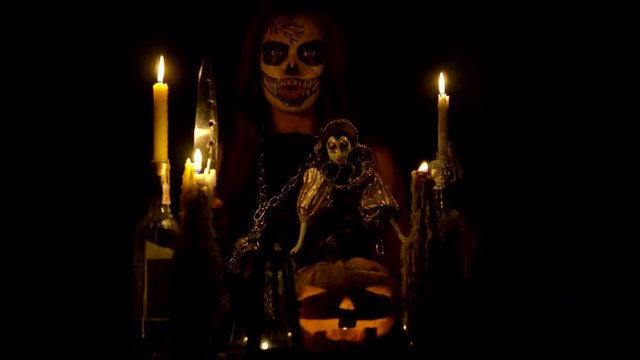 Halloween witch with skull makeup makes voodoo holds knife and wispering spell magic pumpkin chains and candles