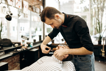 Fashion barber with mustache dressed in a black shirt with a red bow tie tidies up men's beard ...