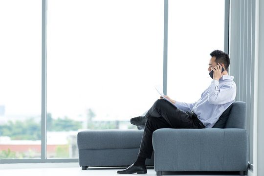 Young Asian businessman using mobile smartphone sitting on sofa serious and have  ducuments at hand in the modern office with big windows