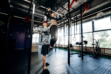 Young athletic man helps slender pretty girl to do pull up on the bar in the gym