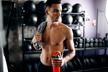 Fototapeta na wymiar Athletic man with a naked torso keeps t-shirt on his shoulder and holds bottle with water in the hand near the sport equipment in the gym