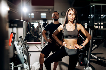 Fototapeta na wymiar Slim dark-haired girl dressed in black sport clothes and fitness coach are doing warm up in the gym