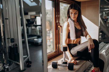 Beautiful athletic girl dressed in black sports top and tights is sitting on the windowsill with  dumbbells next to her