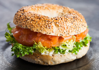 Fresh healthy bagel sandwich with salmon, ricotta and lettuce in black plate on dark kitchen table background. Healthy diet food.
