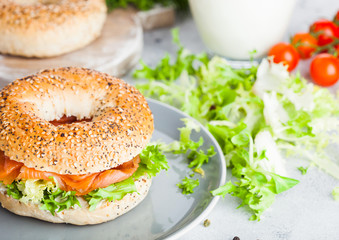 Fresh healthy bagel sandwich with salmon, ricotta and lettuce in grey plate on light kitchen table background. Healthy diet food. Glass of milk and fresh vegetables