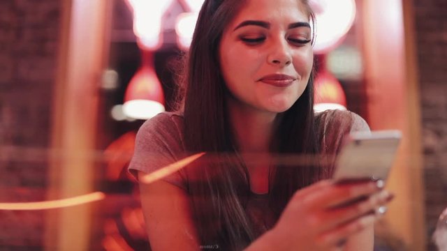 Young woman sitting in bar, drinking lemanade and using her smart phone next to neon bar sign. Woman enjoying night-life while communicating with friends on her smartphone