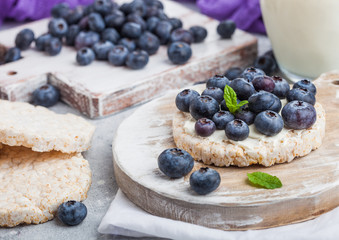 Healthy organic rice cakes with ricotta and fresh blueberries on wooden board and glass of milk on light stone kitchen background. Top view.