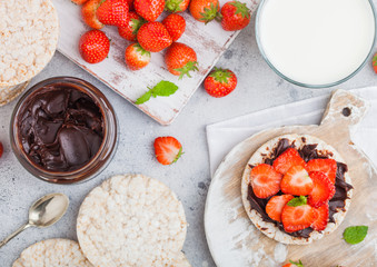 Healthy organic rice cakes with chocolate butter and fresh strawberries on wooden board and glass of milk on light stone kitchen background. Top view.