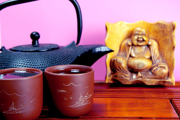 Metal black teapot, two cups of tea on the traditional wooden tea table. Hotei statuette on the background. Pink background.