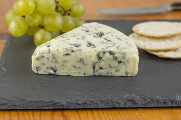 Blue cheese wedge on slate board close up with defocused green grapes and crackers in background