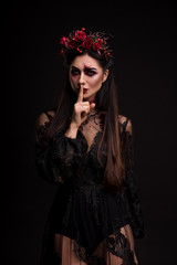 Portrait of a girl in the image for Halloween. Black Widow. Wreath of roses. Finger on lips. Makeup for halloween. Eyes like gimlets. Black lace dress. Black background.