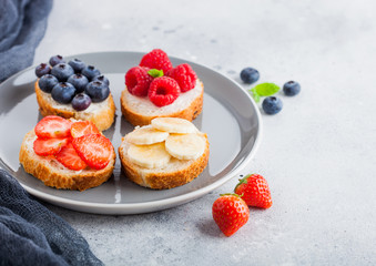 Fresh healthy mini sandwiches with cream cheese, fruits and berries in grey plate with cloth. Strawberries, blueberries, bananas and raspberries on stone kitchen table background.Top view.