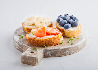 Fresh healthy mini sandwiches with cream cheese, fruits and berries on vintage wooden board. Strawberries, blueberries, bananas and raspberries on stone kitchen table background.Top view.