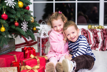 Fototapeta na wymiar Two small children, brother and sister, are sitting on the background of the Christmas interior and gifts. Merry Christmas and Happy Holidays! Christmas tree indoors.