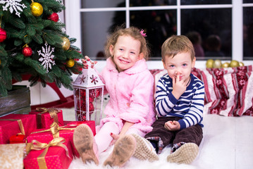 Fototapeta na wymiar Two small children, brother and sister, are sitting on the background of the Christmas interior and gifts. Merry Christmas and Happy Holidays! Christmas tree indoors.