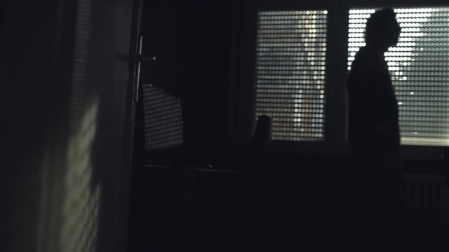 Depressive man in dark room standing by the window, defocussed person in the background