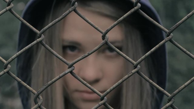 Portrait of moody and sad blonde caucasian girl behind the iron fence. Young woman behind metal fence grid jail locked