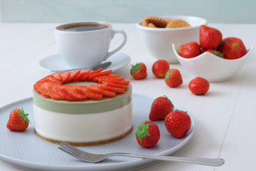 Cooking demo. No-bake two-layered mini strawberry matcha cheesecake. Strawberry slices decorate the top of the cake. 3 cups with coffee, cookies and strawberry in background.