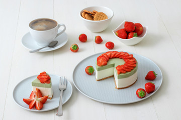 Cooking demo. No-bake two-layered mini strawberry matcha cheesecake. Strawberry slices decorate the top of the cake. 3 cups with coffee, cookies and strawberry in background.