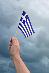 Hand holding Greek flag high in the air, with a stormy, cloudy sky
