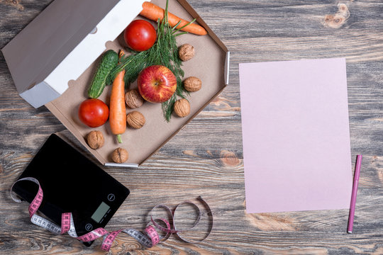 Weight loss and healthy eating concept. Open pizza box with raw vegetables in it, kitchen scales and measuring tape and paper on wooden background. Top view, flat lay.