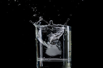 Bounce and splash from a piece of ice thrown into a transparent glass with water on a black background