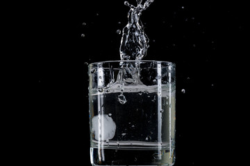 Bounce and splash from a piece of ice thrown into a transparent glass with water on a black background