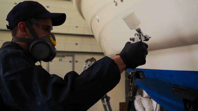 Professional master man in a robe and puller with spray gun puts a layer of paint or ceramics on the yacht. Concept of: Dangerous work, Chemistry, Paints, Garage, Transport Service.