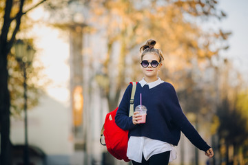 Naklejka premium Pretty hipster teen with red bag drinks milkshake from a plastic cup walking street between buildings. Cute girl in sunglasses drinks a drink through a straw.
