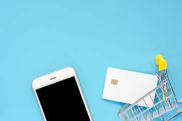 Smartphone, blank white credit card and mini shopping cart or trolley on blue background for business and technology concept