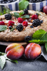 Whole-grain galette with plums and berries on dark background, top view