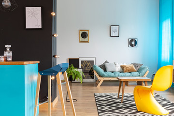 Modern yellow chair in eclectic living room with blue kitchen island, black wall and comfortable sofa with posters on the empty white wall